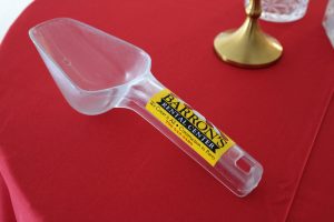an opaque ice scoop with a yellow barrons rental center sticker on the handle. the scoop sits on a red tablecloth.