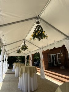 Inside a white marquee tent with three chandeliers that contain lemons. Three pedestal tables sit in the center lined up, with a long white table cloth and a smaller yellow tablecloth topper on each one.